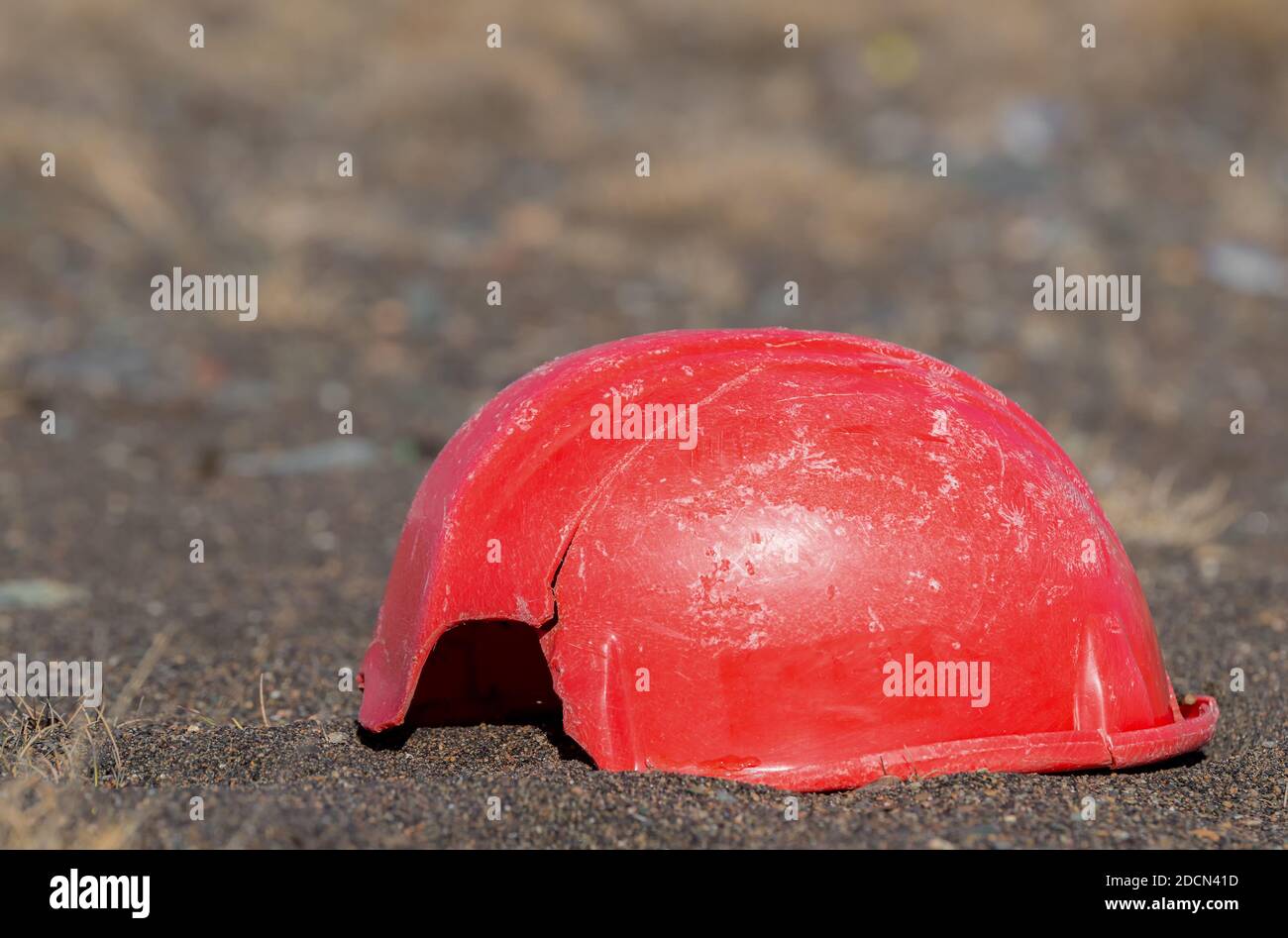 An old, broken hardhat lying on the ground. It is a faded and scratched red, and has a piece missing from it. It is also cracked. There is room for te Stock Photo