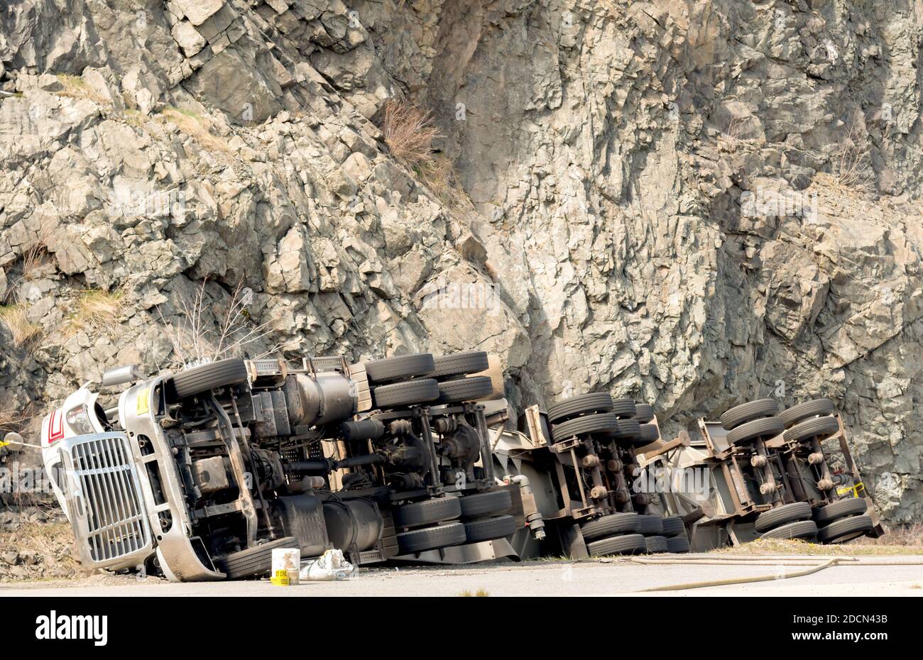 A large tanker truck flipped on its side in a ditch. The truck has 30 wheels, and the front appears damaged. A high cliff is in the background. Stock Photo