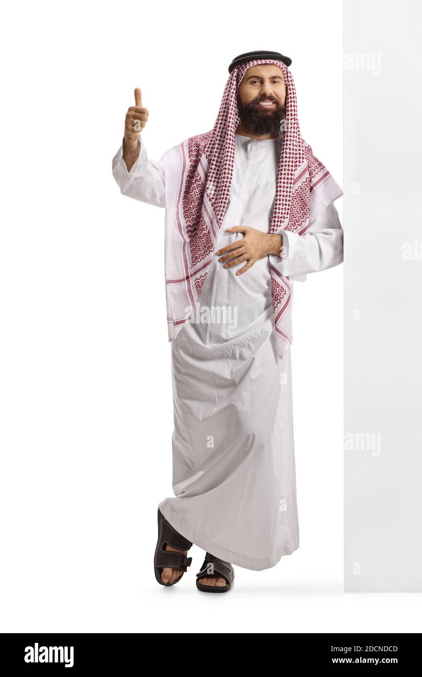 Full length portrait of a saudi arab man wearing a traditional thobe, leaning on a wall and gesturing thumbs up isolated on white background Stock Photo