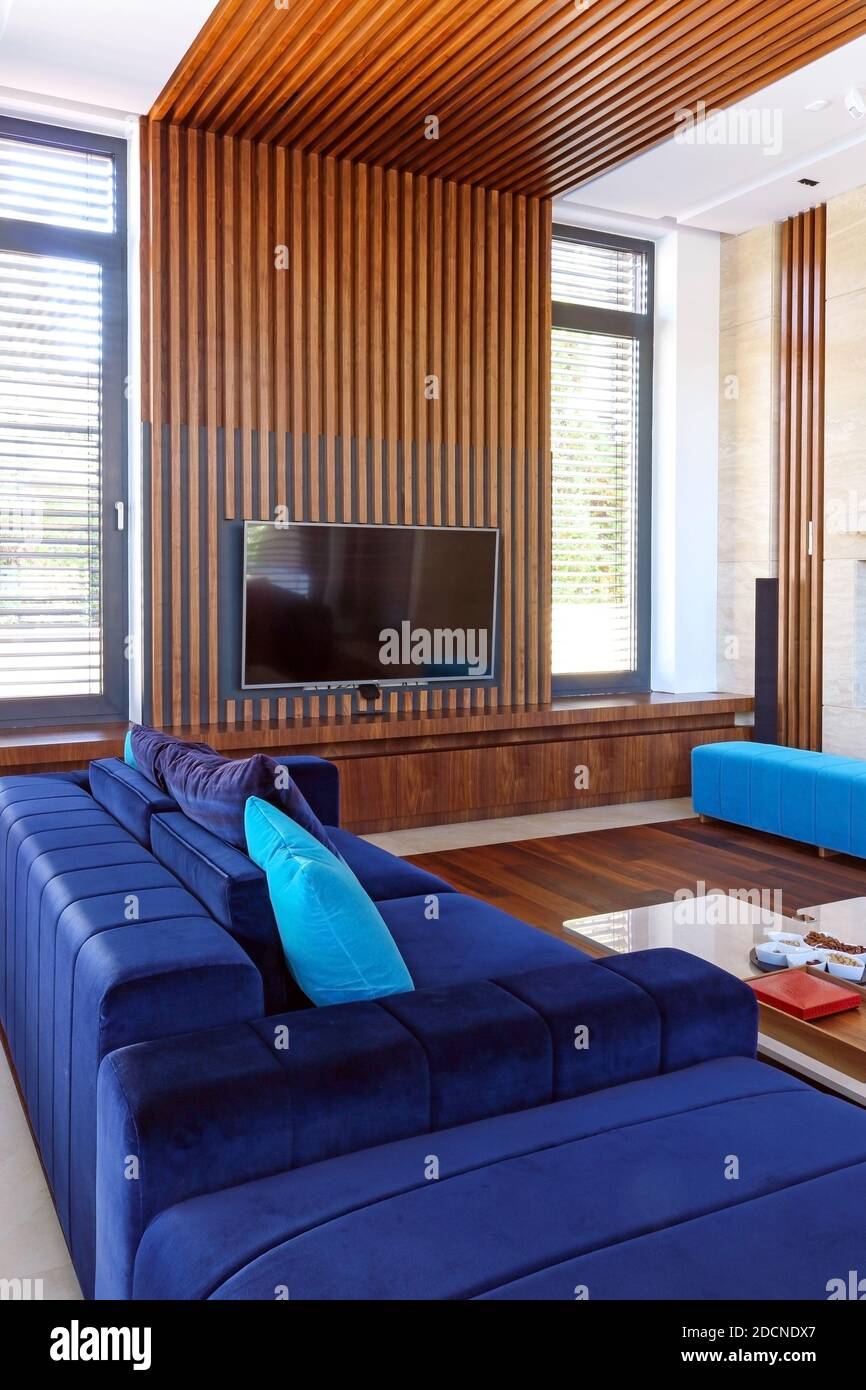 Modern interior with wood slat  ceiling, comfortable blue furniture, parquet, white table and wooden tv wall unit. Living room with big windows and wo Stock Photo