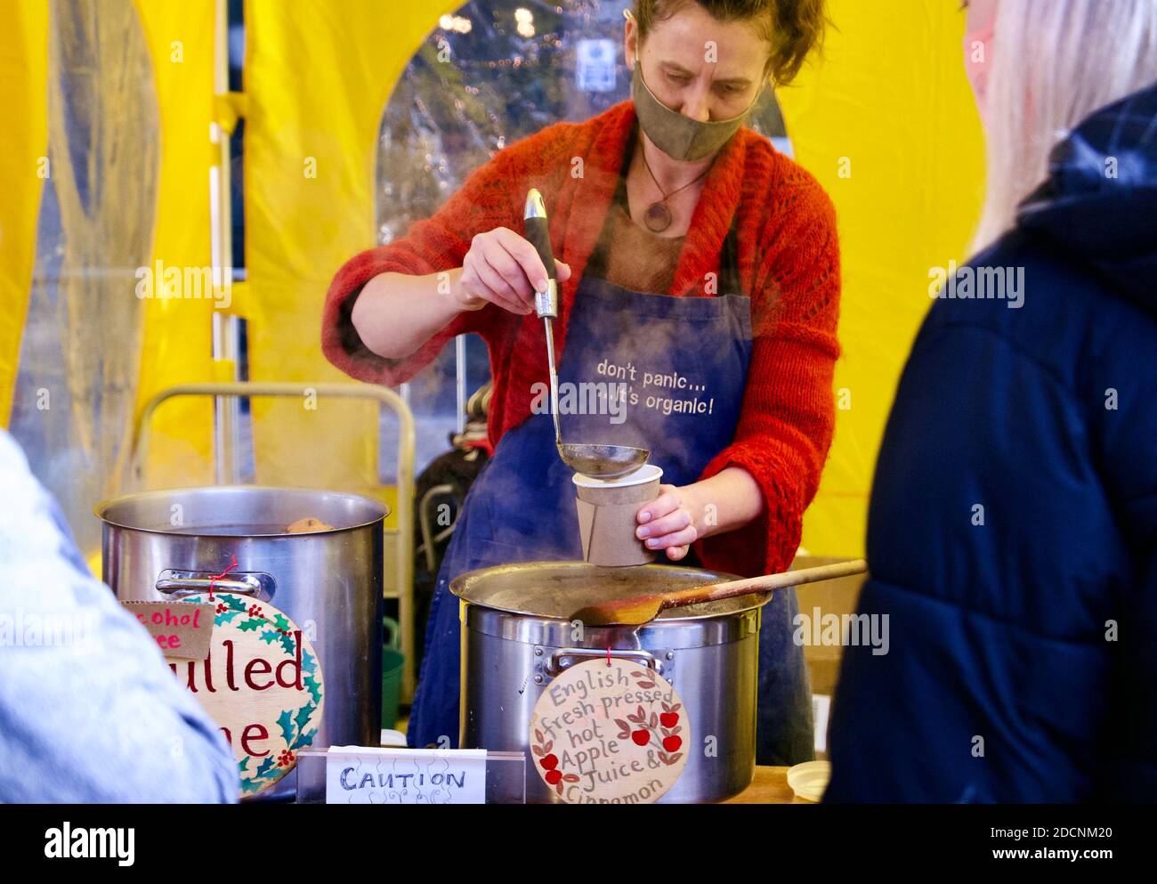 Woman wearing a face mask serves hot apple juice and cinnamon at a Christmas food market stall in Coal Drops Yard, Kings Cross, London. Stock Photo