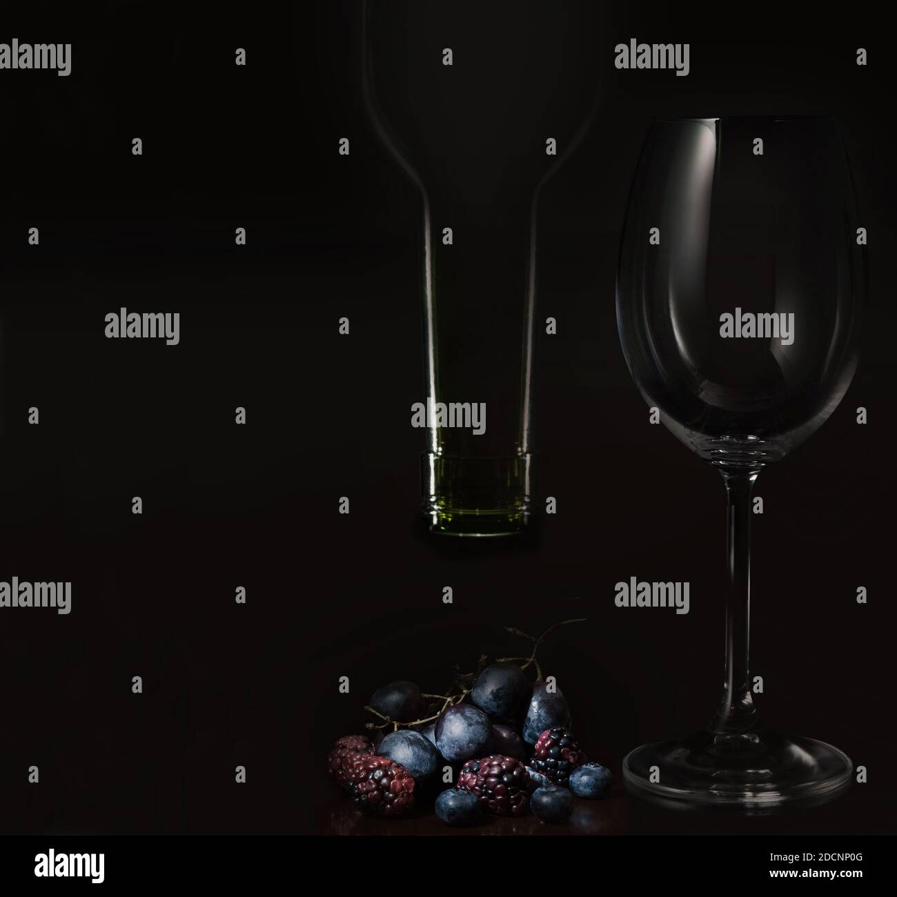 The neck of the wine bottle (upside down), ripe forest fruits and  grapes and an empty wine glass, all in an atmosphere of chiaroscuro.. Stock Photo