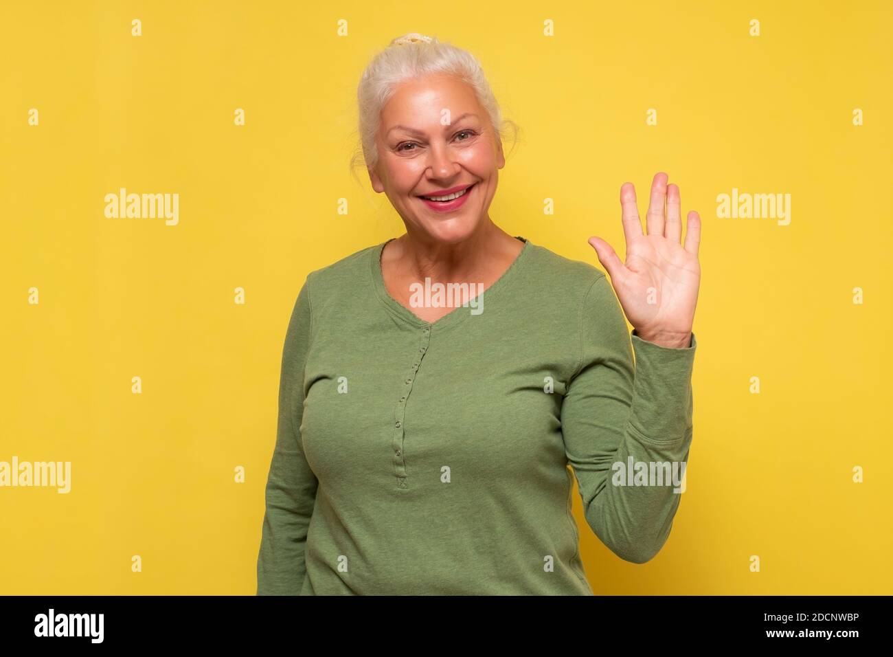 Senior friendly looking woman smiling happily, saying Hello, Hi or Bye, waving hand at camera. Stock Photo