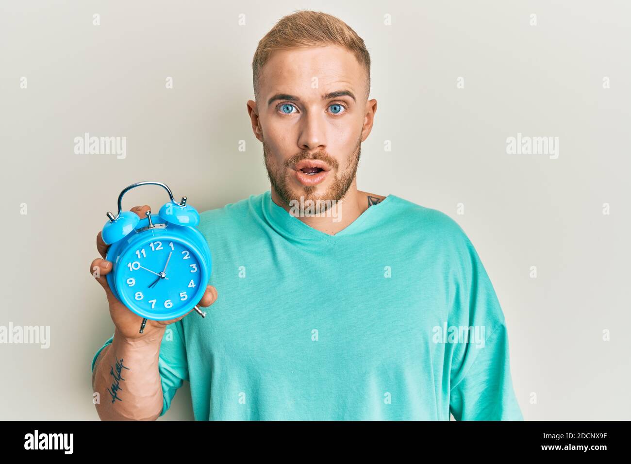 Young caucasian man holding alarm clock scared and amazed with open