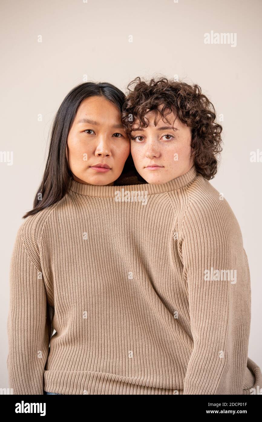 Young Asian and Caucasian women with dark hair in one knitted beige sweater Stock Photo