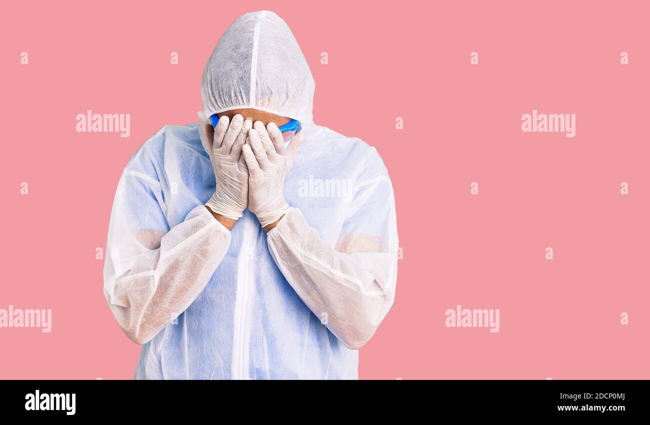 Young hispanic man wearing doctor protection coronavirus uniform and medical mask with sad expression covering face with hands while crying. depressio Stock Photo