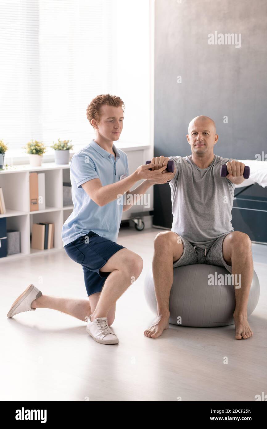 Young physiotherapist helping male patient to do exercise with dumbbells Stock Photo