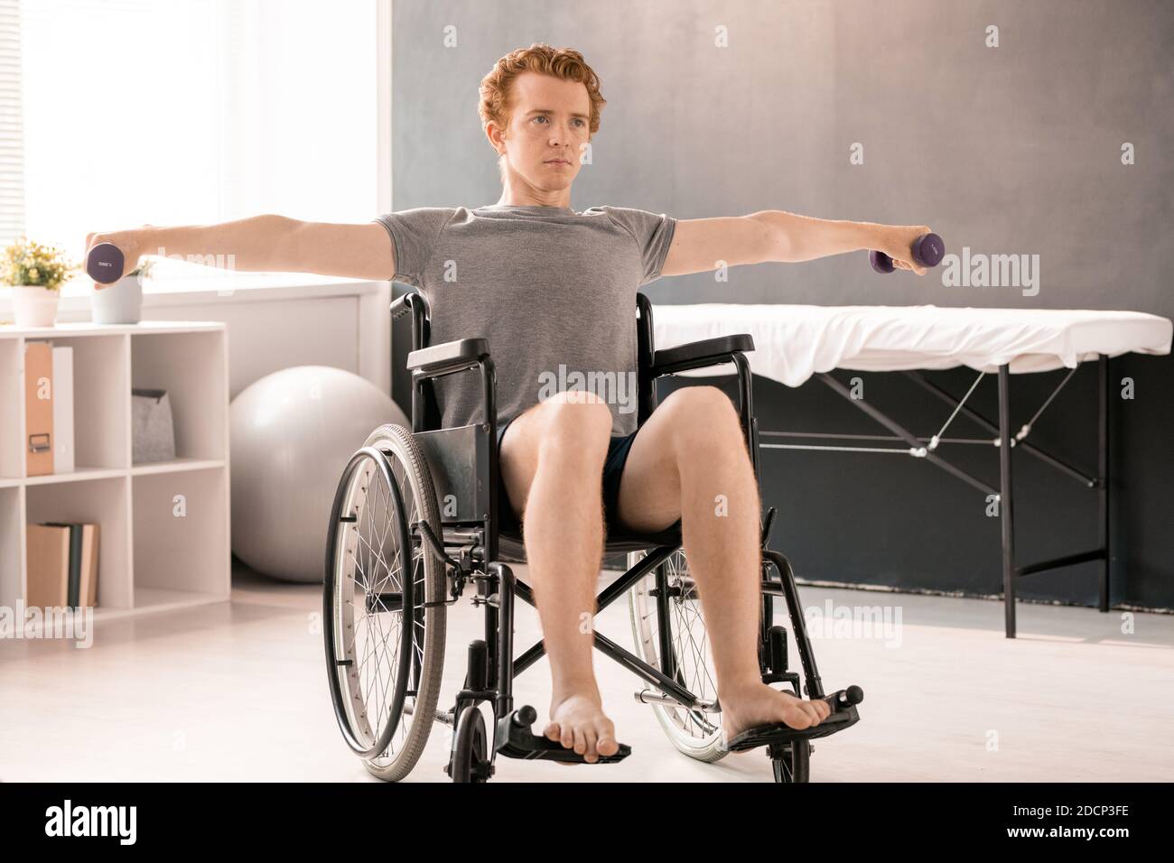 Young sportsman sitting in wheelchair and outstretching arms with dumbbells Stock Photo
