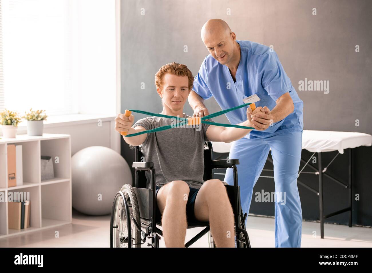 Mature clinician in blue uniform supporting hand of young patient in wheelchair Stock Photo