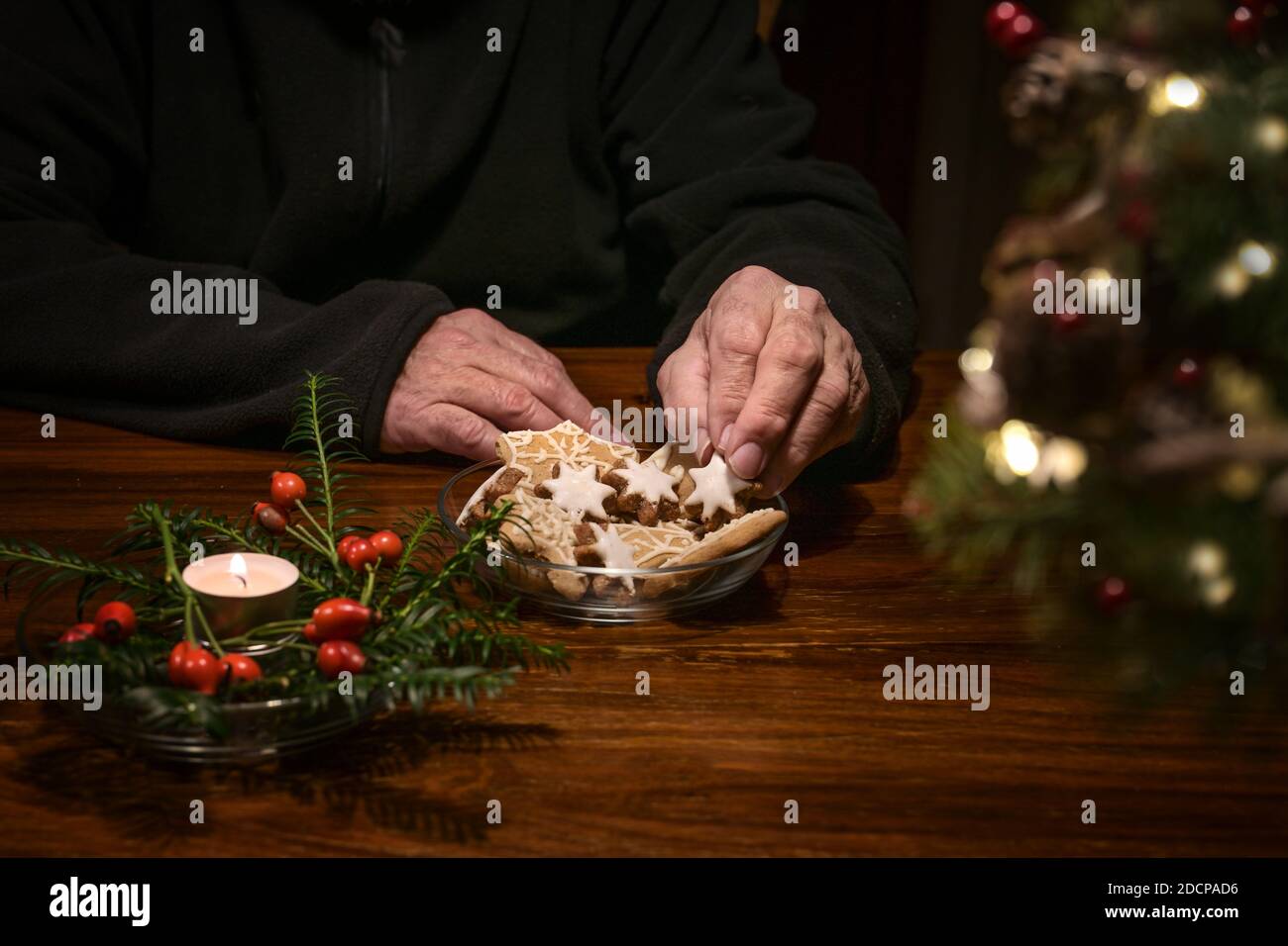 Hands of a lonely old man taking Christmas cookies on a table with burning candle and festive decoration, sad holidays during the coronavirus pandemic Stock Photo