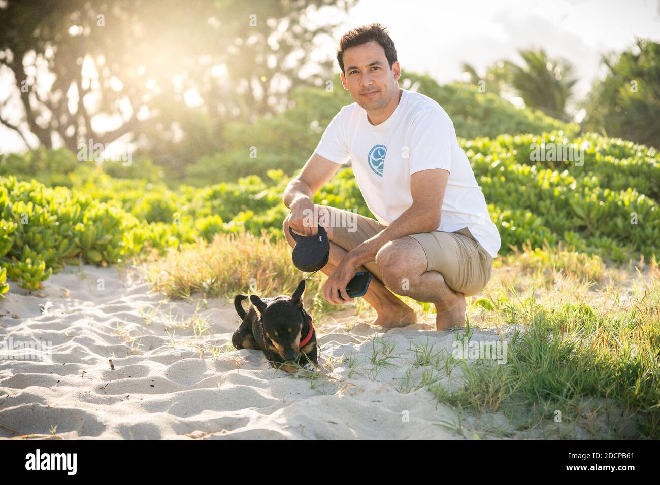 Young fit male squats on the beach with his small black dog in Hawaii Stock Photo