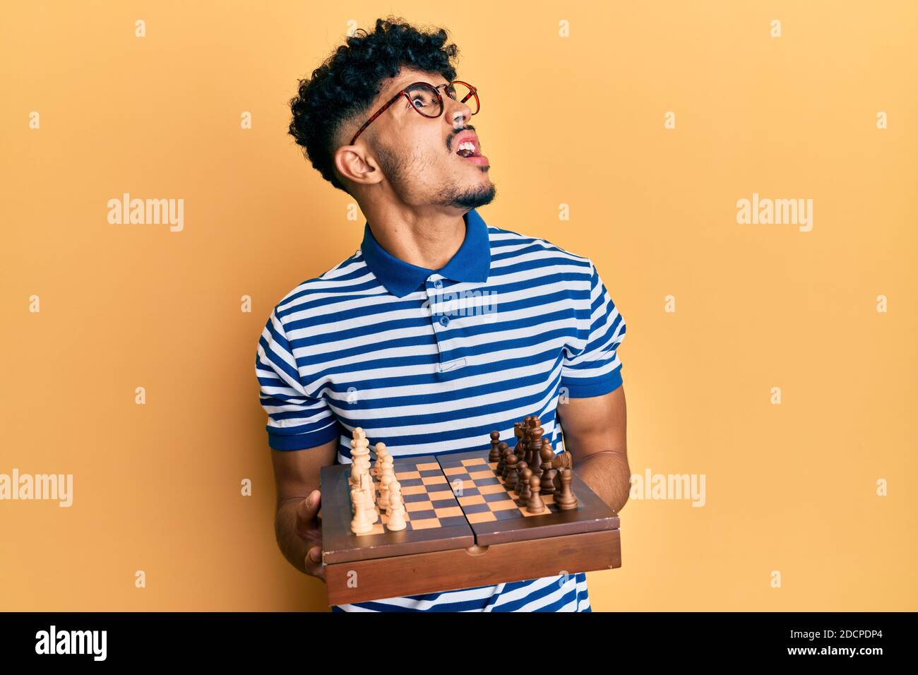 Young arab handsome man holding chess board wearing glasses angry and mad screaming frustrated and furious, shouting with anger looking up. Stock Photo