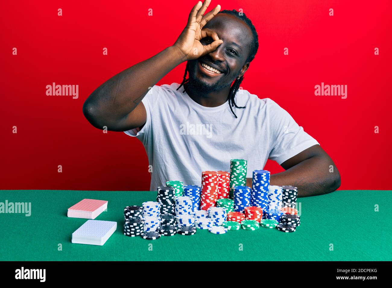young black man sitting on the table with poker chips and cards smiling happy doing ok sign with hand on eye looking through fingers Stock Photo - Alamy