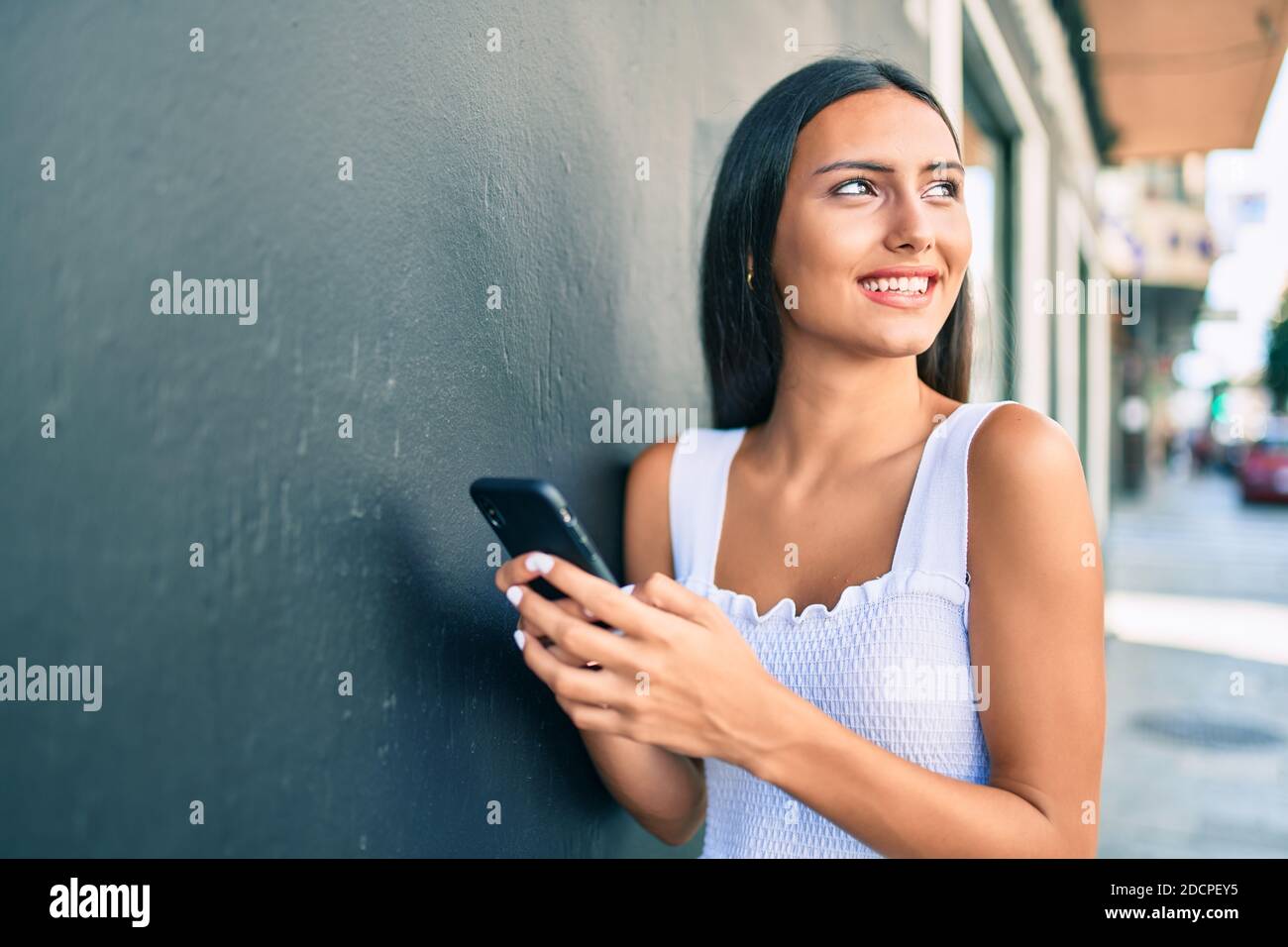 Young latin girl smiling happy using smartphone leaning on the wall ...