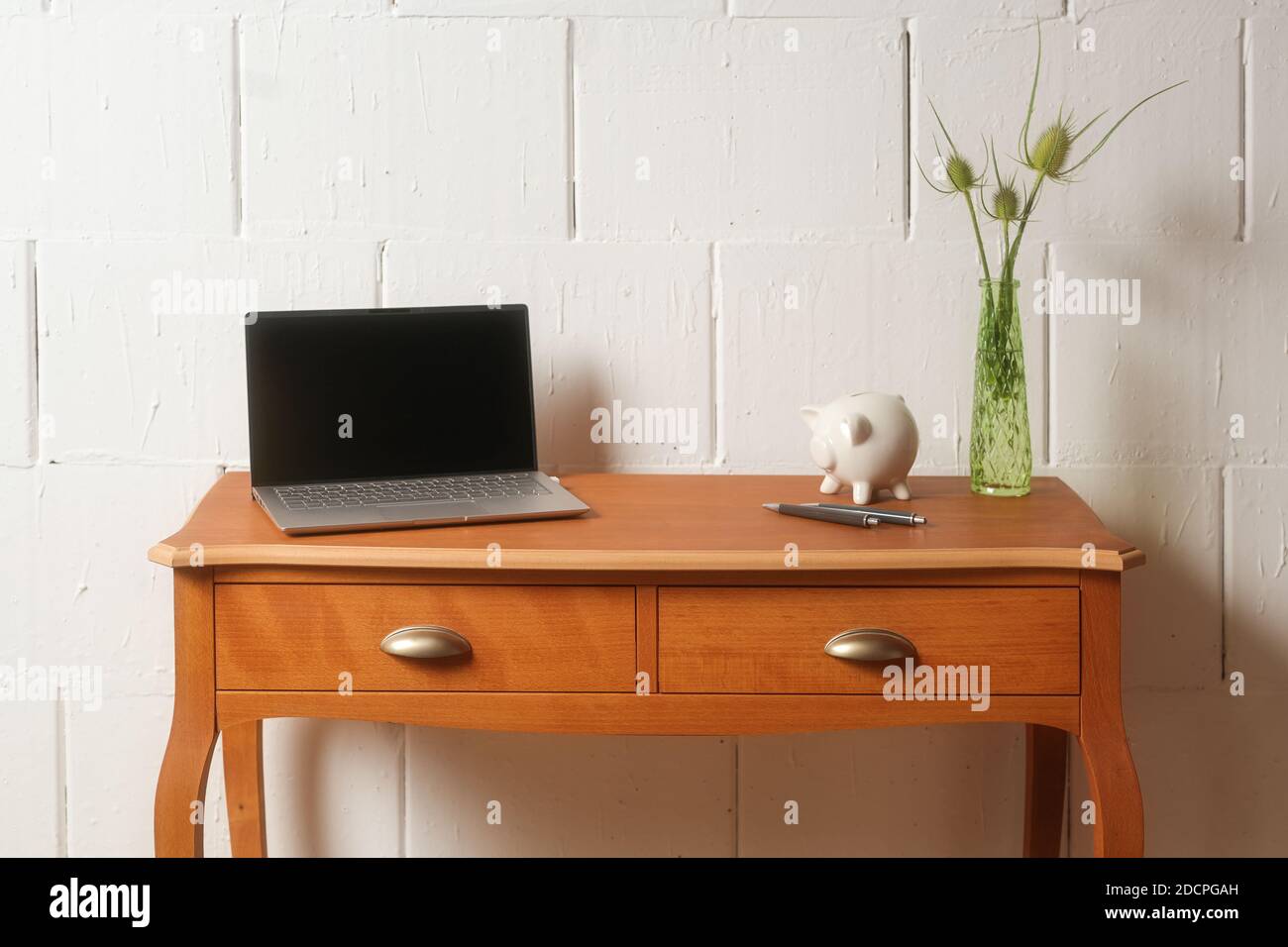 Small wooden vintage table with laptop, piggy bank and flower vase against a rough white wall, tiny home office, copy space, selected soft focus, narr Stock Photo