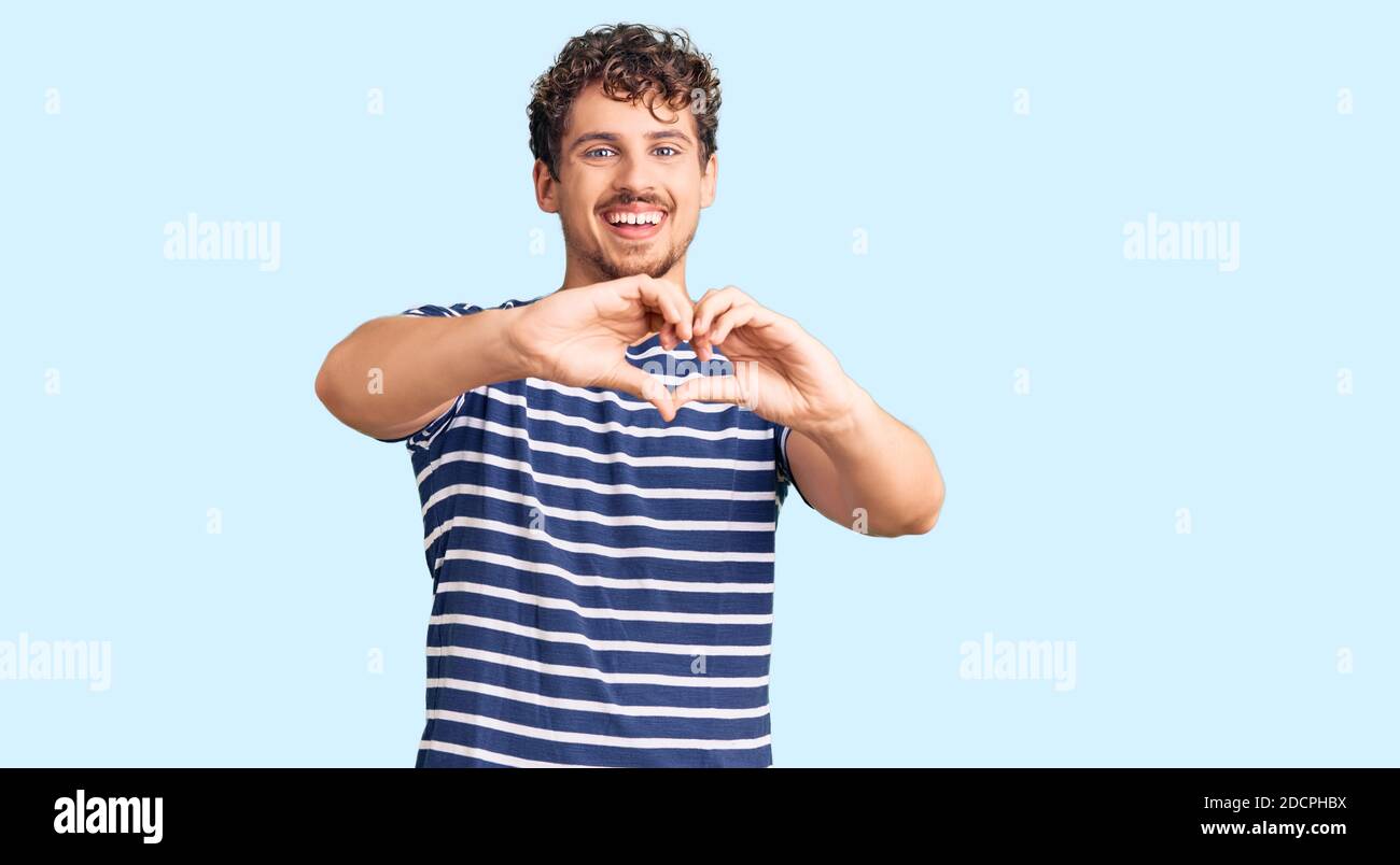 Young handsome man with curly hair wearing casual clothes smiling in love doing heart symbol shape with hands. romantic concept. Stock Photo