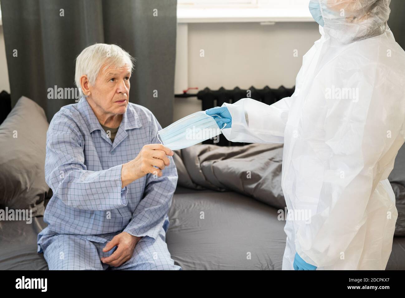 Sick senior man in blue pajamas taking medical mask from gloved hand of doctor Stock Photo
