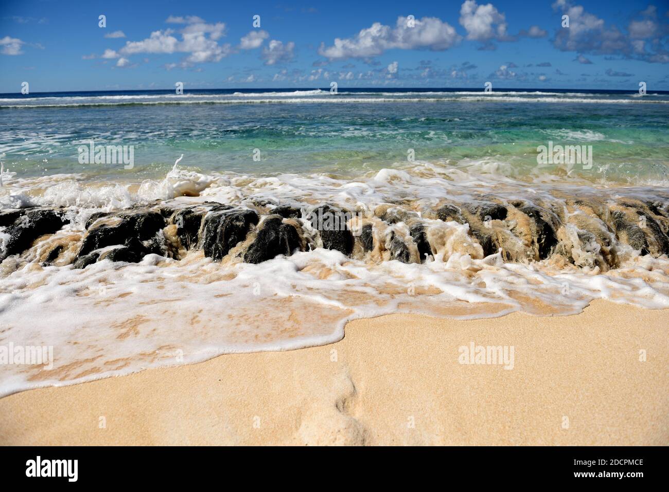 Ocean waves spilling over lava reef rocks, sandy beach, blue sea, sky and clouds in Guam, Micronesia Stock Photo