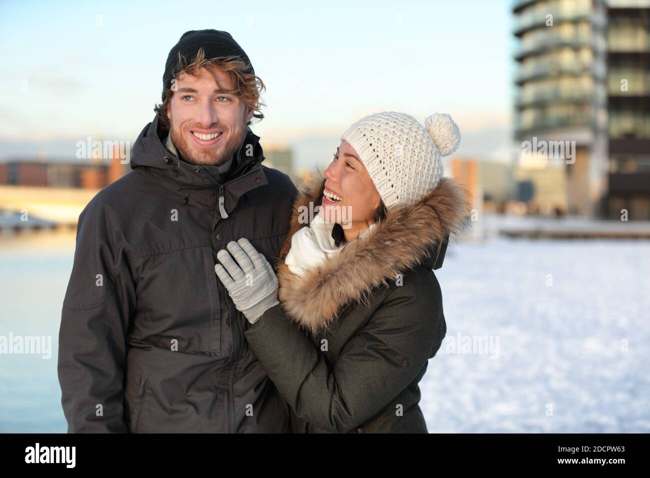 Winter couple in snow hat and coats. Happy young adults walking together smiling on a city street enjoying sunny urban walk in modern neighborhood Stock Photo