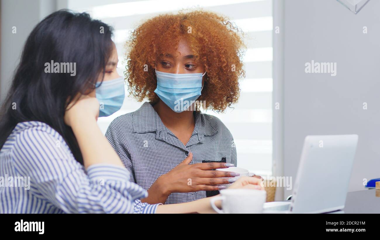 Multiracial workplace and coronavirus protection. Women collegues with medical mask working on the project with coffee. High quality photo Stock Photo