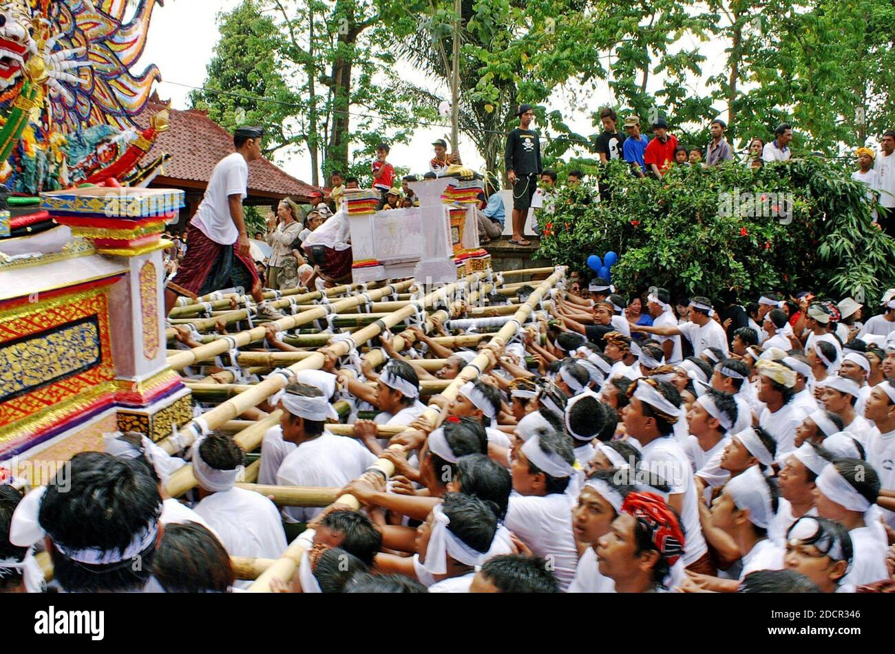 Young Balinese men carry and follow an opulent funeral tower during a procession through Ubud, Bali, Indonesia.  The Balinese Hindu funeral is culturally rich with ritual and custom.  The size and expense of the multi-level casket is based on the caste, wealth, and prestige of the person who passed on.  This massive funeral ceremony and celebration indicates the person was important in Balinese society.  Onlookers watch the procession celebrating life, death, and reincarnation reflecting the Balinese life-cycle. Stock Photo