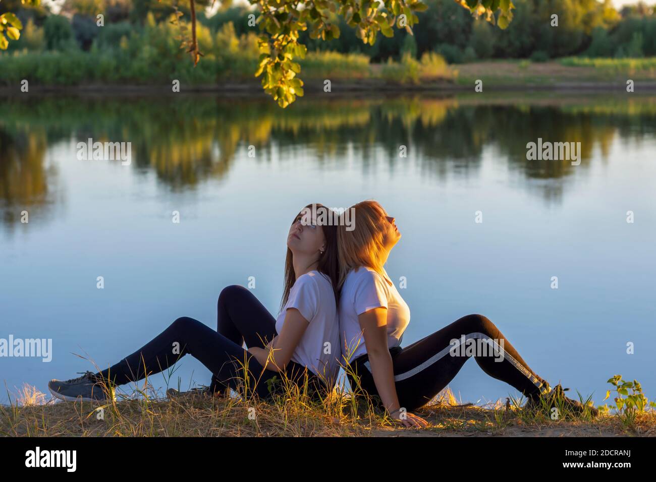 Two athletic young women are resting after training on the river bank. Blonde and the brunette are back to back in nature. Soft focus, blur. Stock Photo