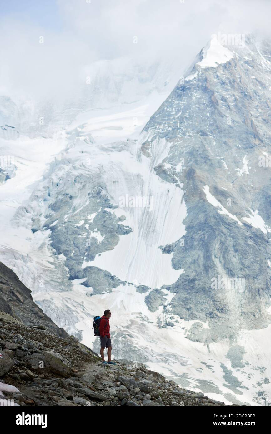 Vertical snapshot of incredibly beautiful mount in Swiss Alps - Ober Gabelhorn, man standing on a slope enjoying a cold quiete scenery. Concept of freedom, mountain hiking, tourism and alpinism Stock Photo