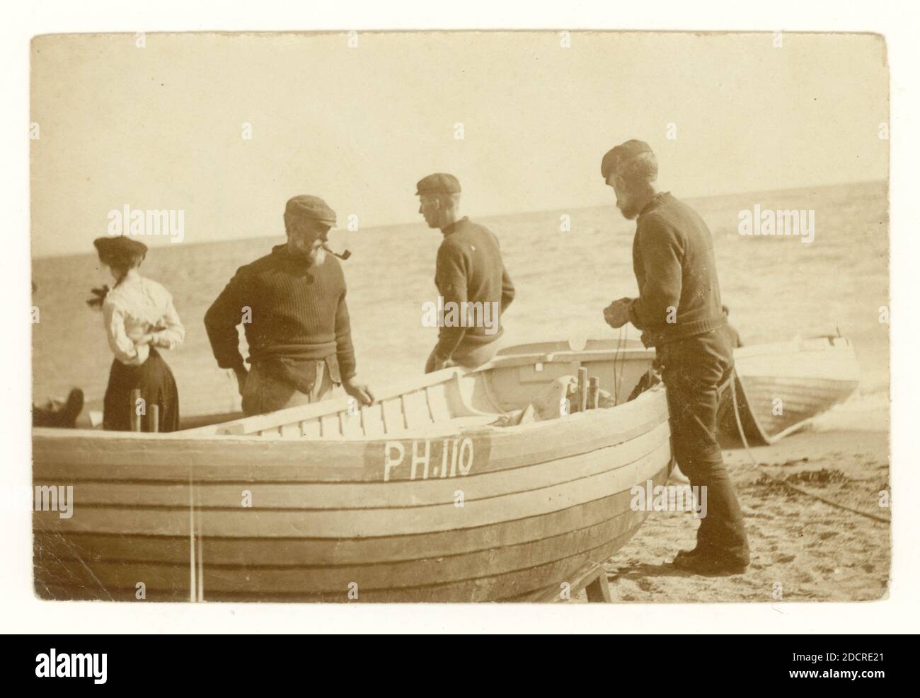 Original early 1900's sepia toned photograph group of fishermen beside their boats which are pulled up on the shore, preparing to go to sea, possibly line fishing, registered in Plymouth as PH 110,  Devon, U.K. circa 1910 Stock Photo