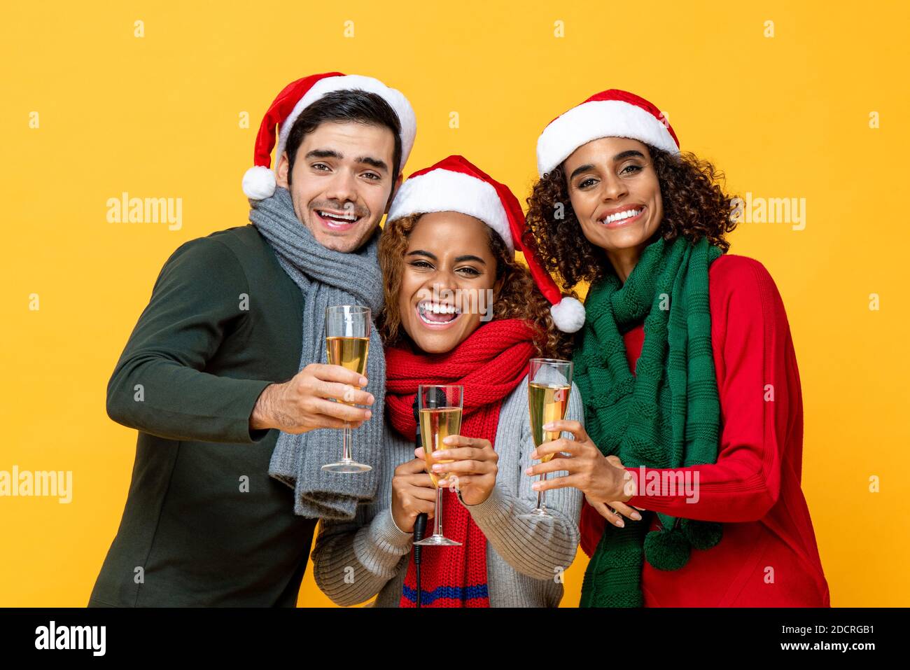 Happy portrait of three diverse friends in Christmas attire clinking glasses of champagne celebrating in studio yellow isolated background Stock Photo