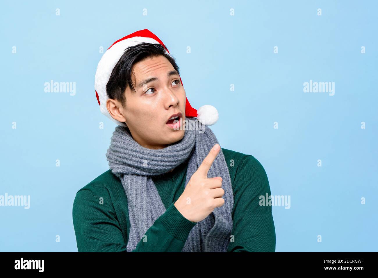 Surprised Asian man in Christmas attire looking and pointing hand up to empty space aside on light blue isolated background Stock Photo