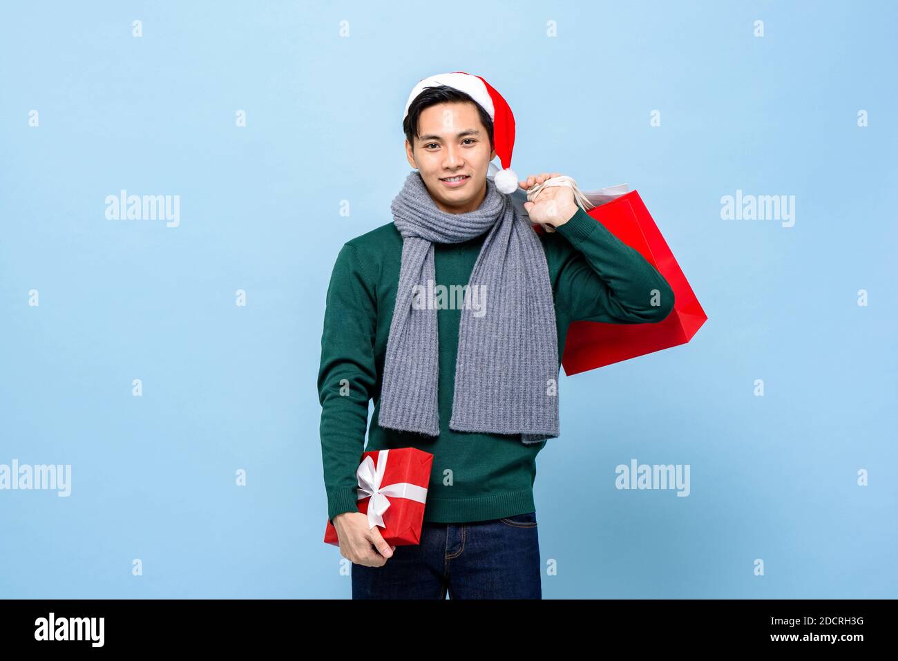 Happy portrait of smiling young handsome Asian man wearing green christmas sweater and red hat holding shopping bags isolated on light blue background Stock Photo