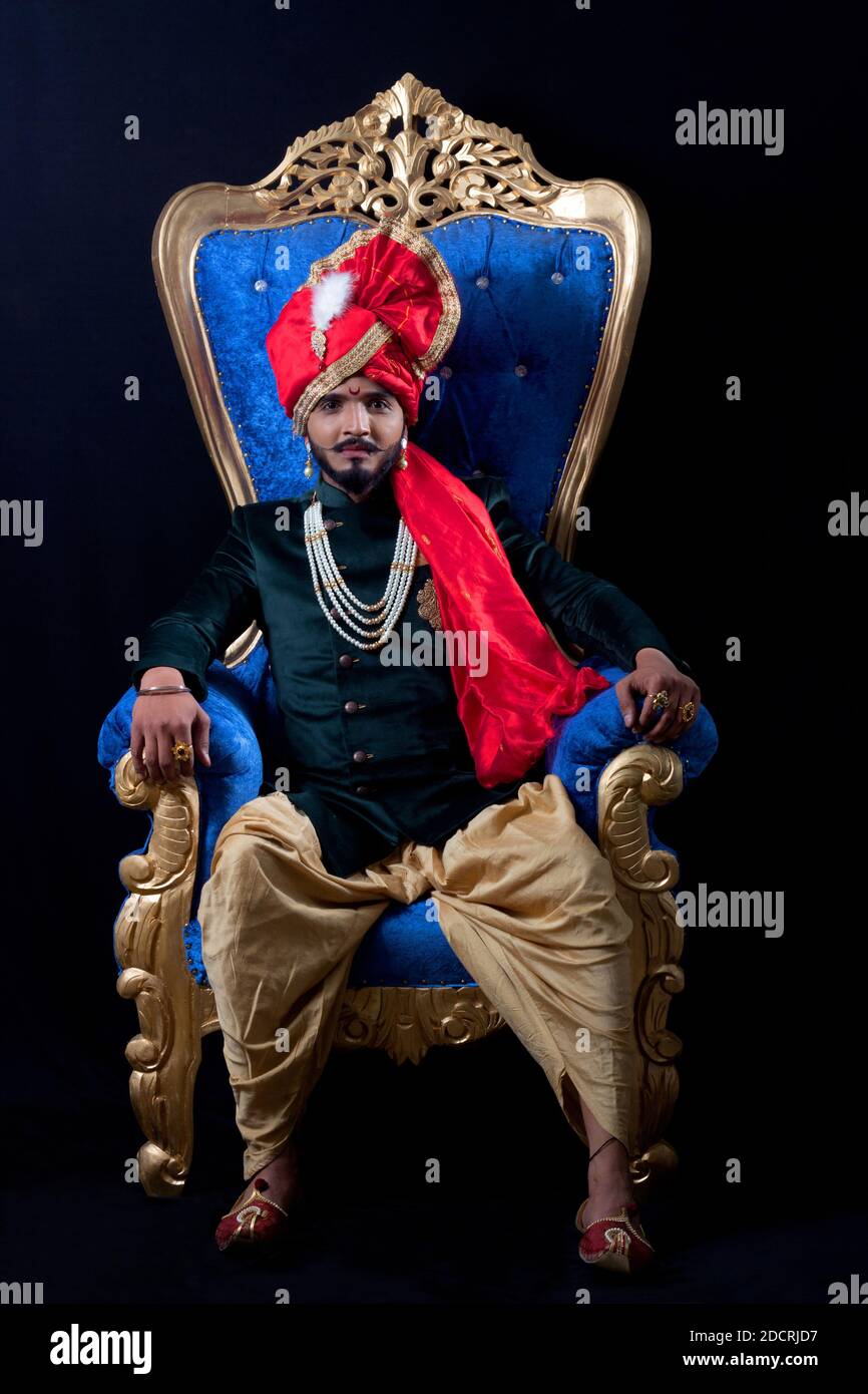 Young bearded man in traditional Indian attire with Dhoti and red turban on  a blue Maharaja chair. Black background Stock Photo - Alamy