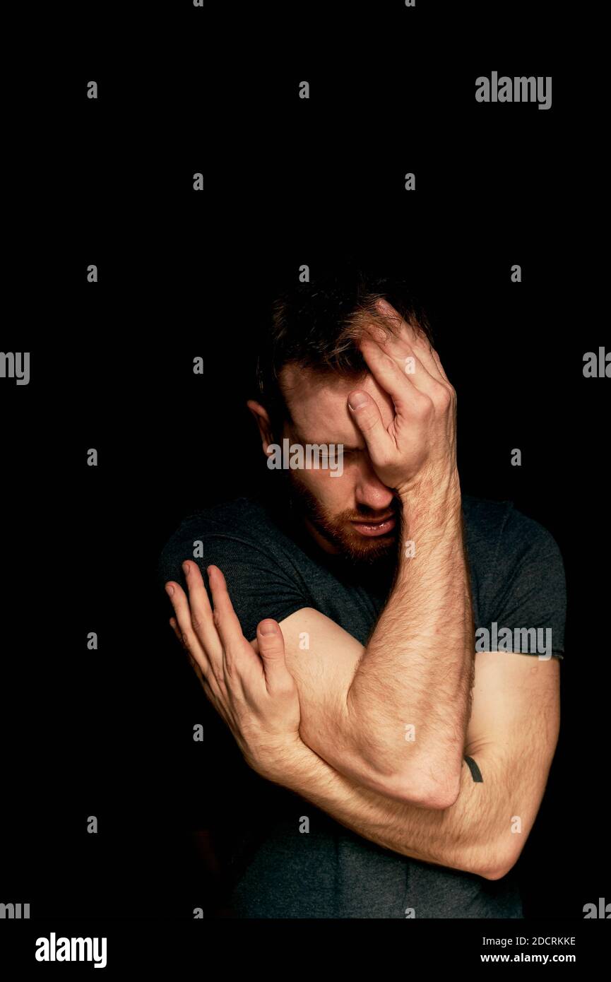 A man on a black background covers his face with his hands. The atmosphere of hopelessness,dejection Stock Photo