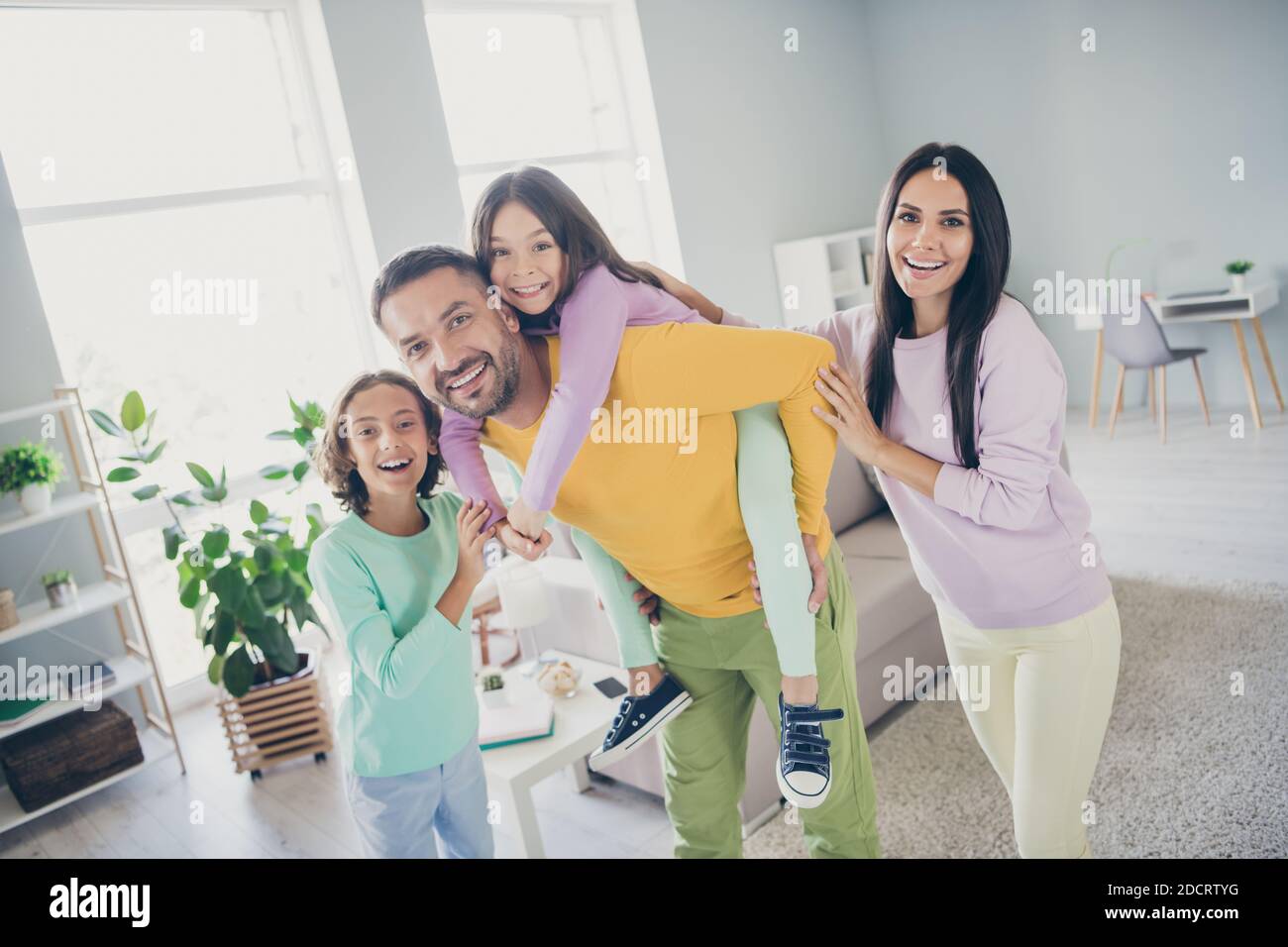 Photo of full family four members dad hold piggy back daughter have fun beaming smile wear colorful pullover in living room indoors Stock Photo