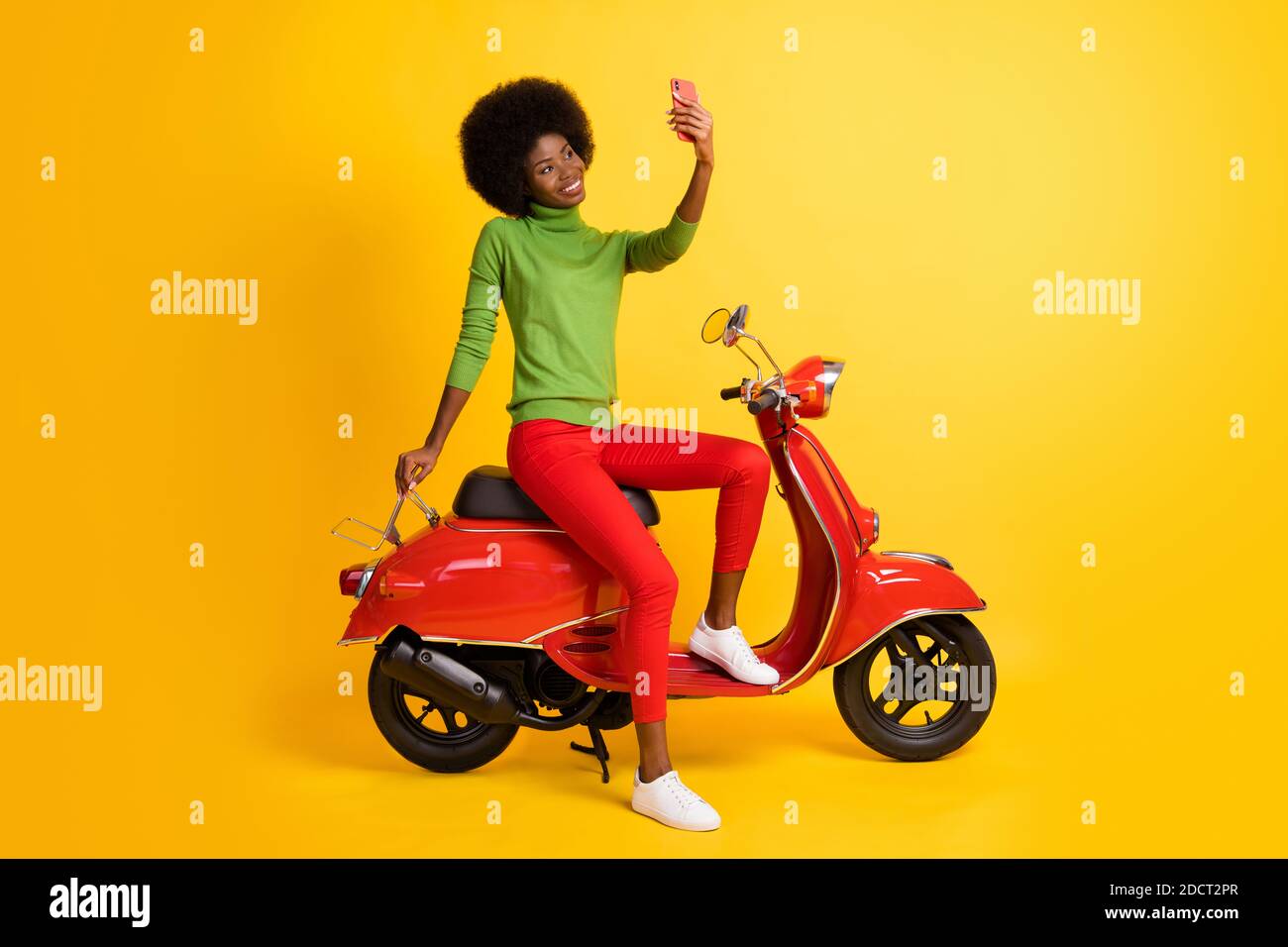 Photo portrait of young rider biker with brunette sitting on red retro motorbike holding cellphone taking selfie isolated on vivid yellow colored Stock Photo