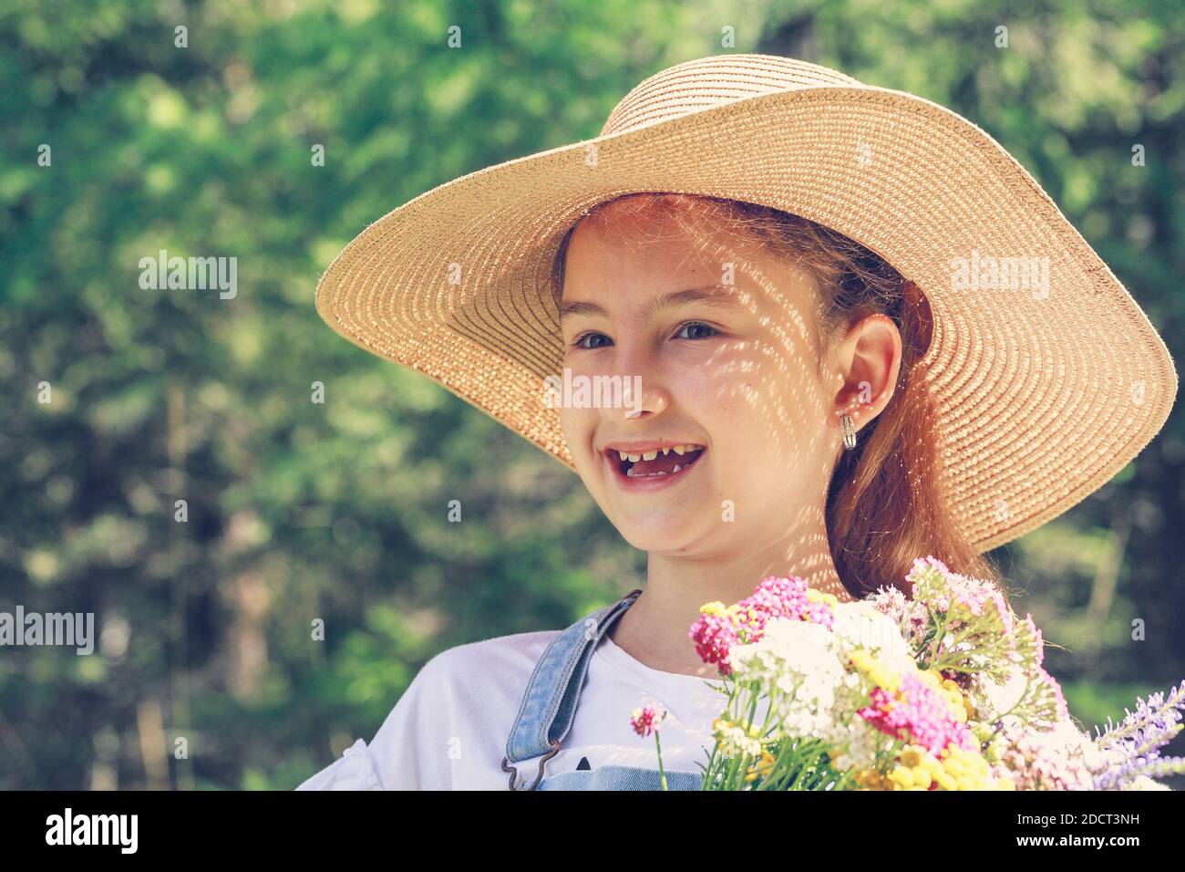 Cute smiling girl with flowers in a straw hat on a summer background. Close-up portrait Stock Photo