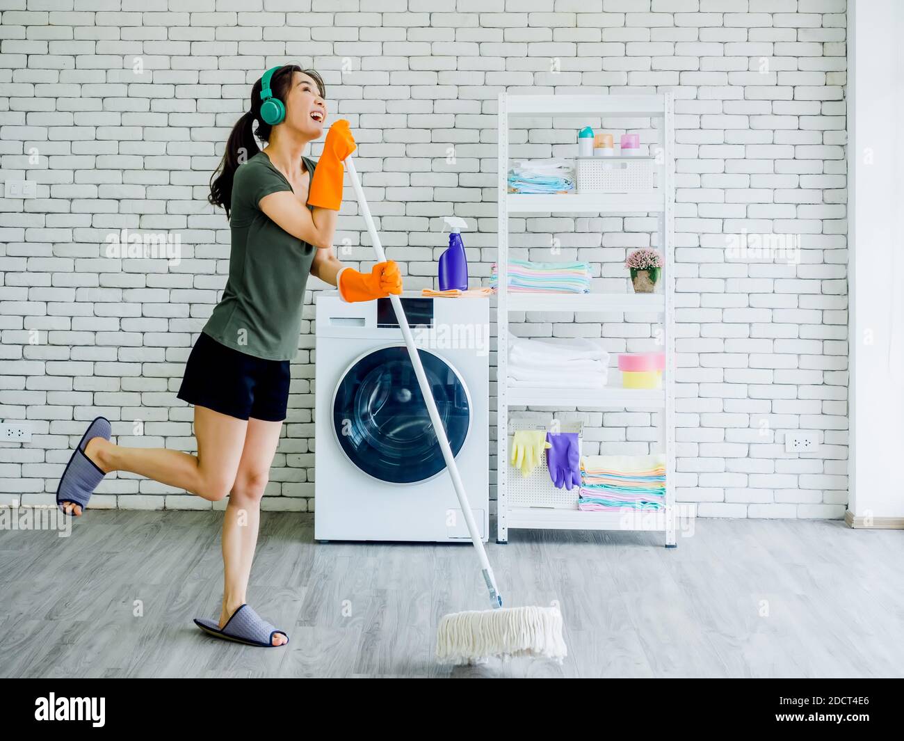 Happy beautiful young Asian woman, housewife wearing rubber gloves, slipper and green headphone singing fun with mop like a microphone while cleaning Stock Photo