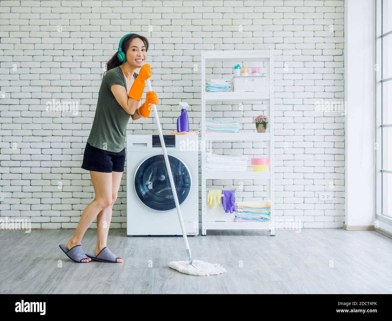 Happy beautiful young Asian woman, housewife wearing rubber gloves, slipper and green headphone singing fun with mop like a microphone while cleaning Stock Photo