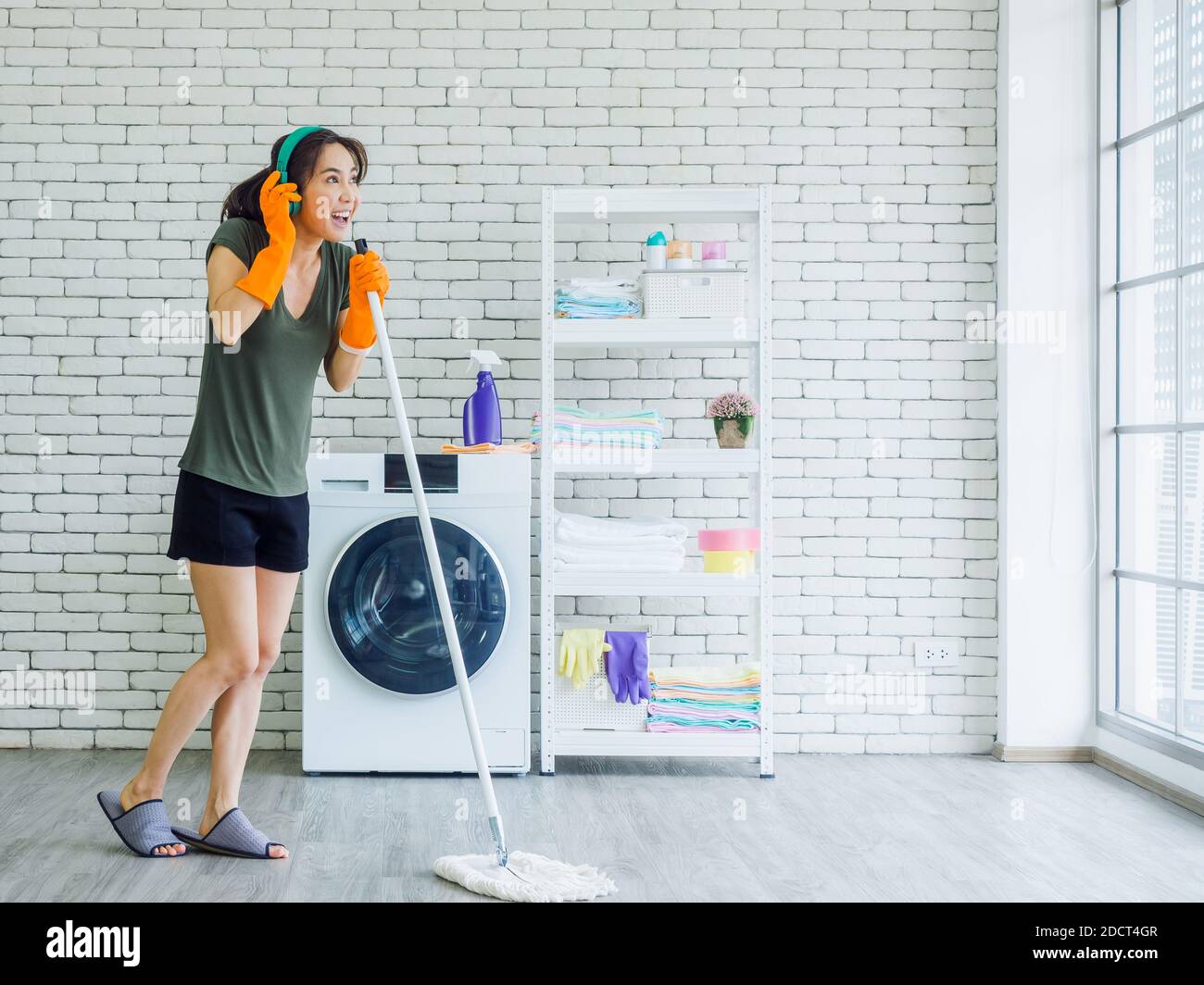 Happy beautiful young Asian woman, housewife wearing rubber gloves, slipper and green headphone singing fun with mop like a microphone while cleaning Stock Photo