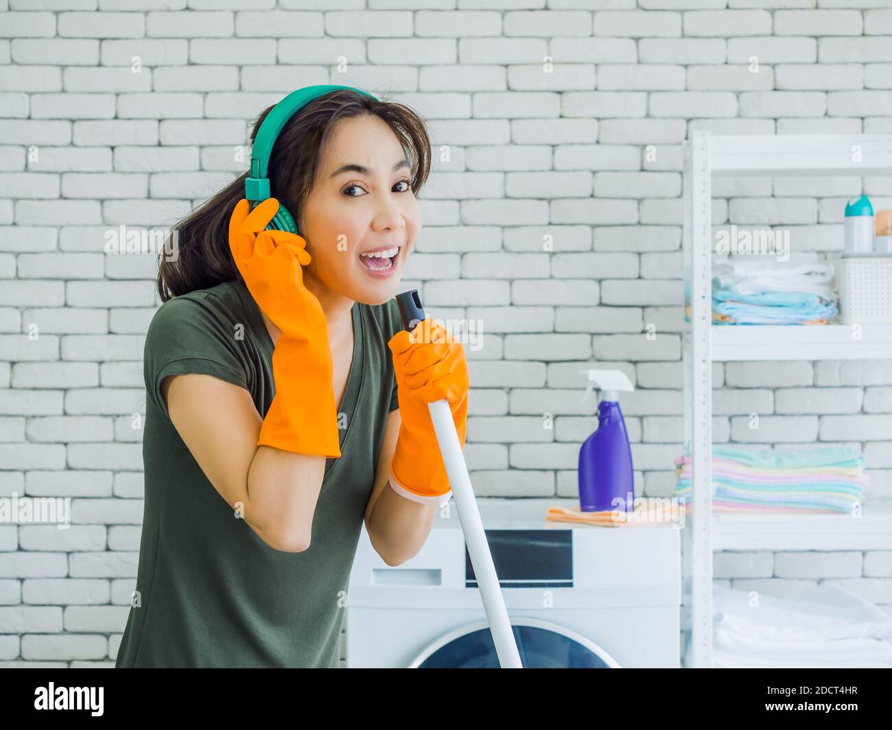 Happy beautiful young Asian woman, housewife wearing orange rubber gloves, listening music with green headphone singing fun with mop like a microphone Stock Photo