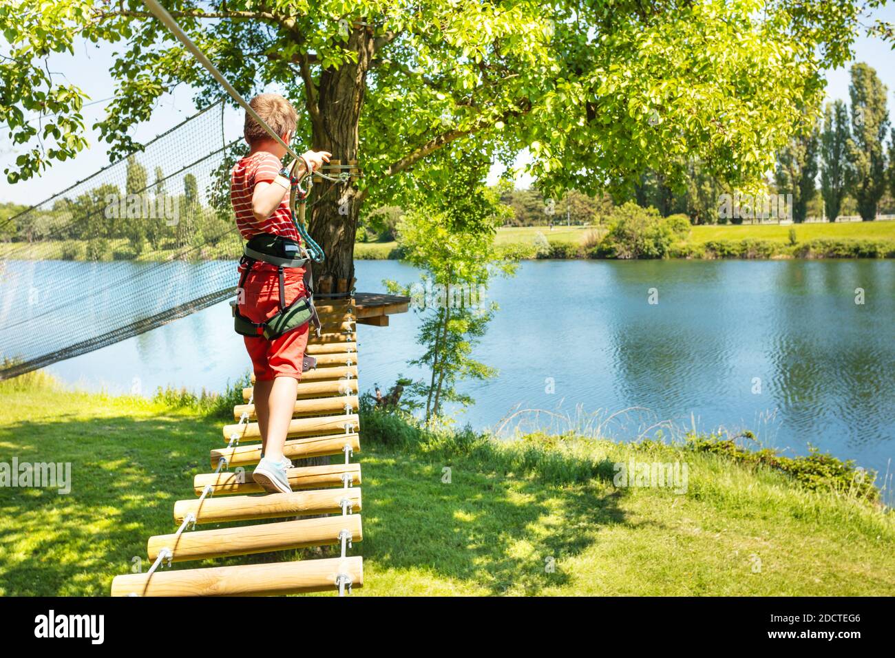 Little boy walk on the sky rope bridge between two trees near the lake in summer amusement camp playground Stock Photo