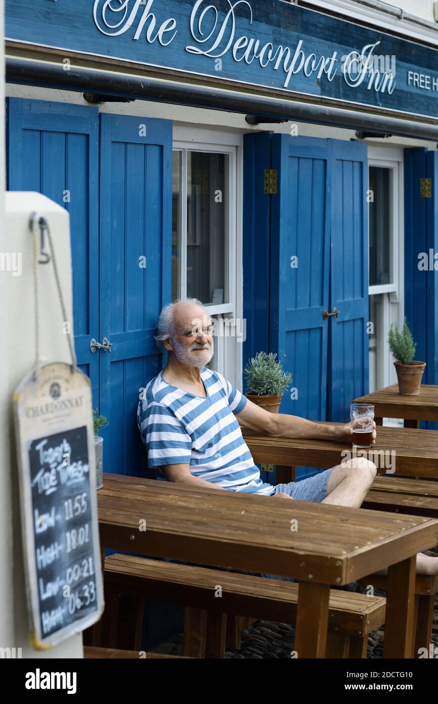 Happy man wearing a breton striped shirt sitting outside a pub drinking a pint in Kingsand ,Cornwall , UK Stock Photo