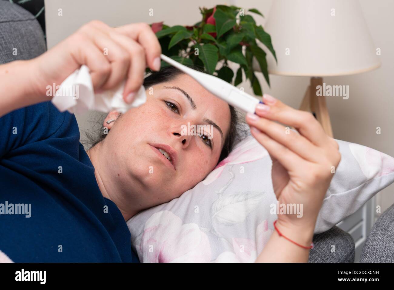 Sick female model with influenza cold covid19 symptoms looking at thermometer checking body temperature high fever feeling unwell Stock Photo