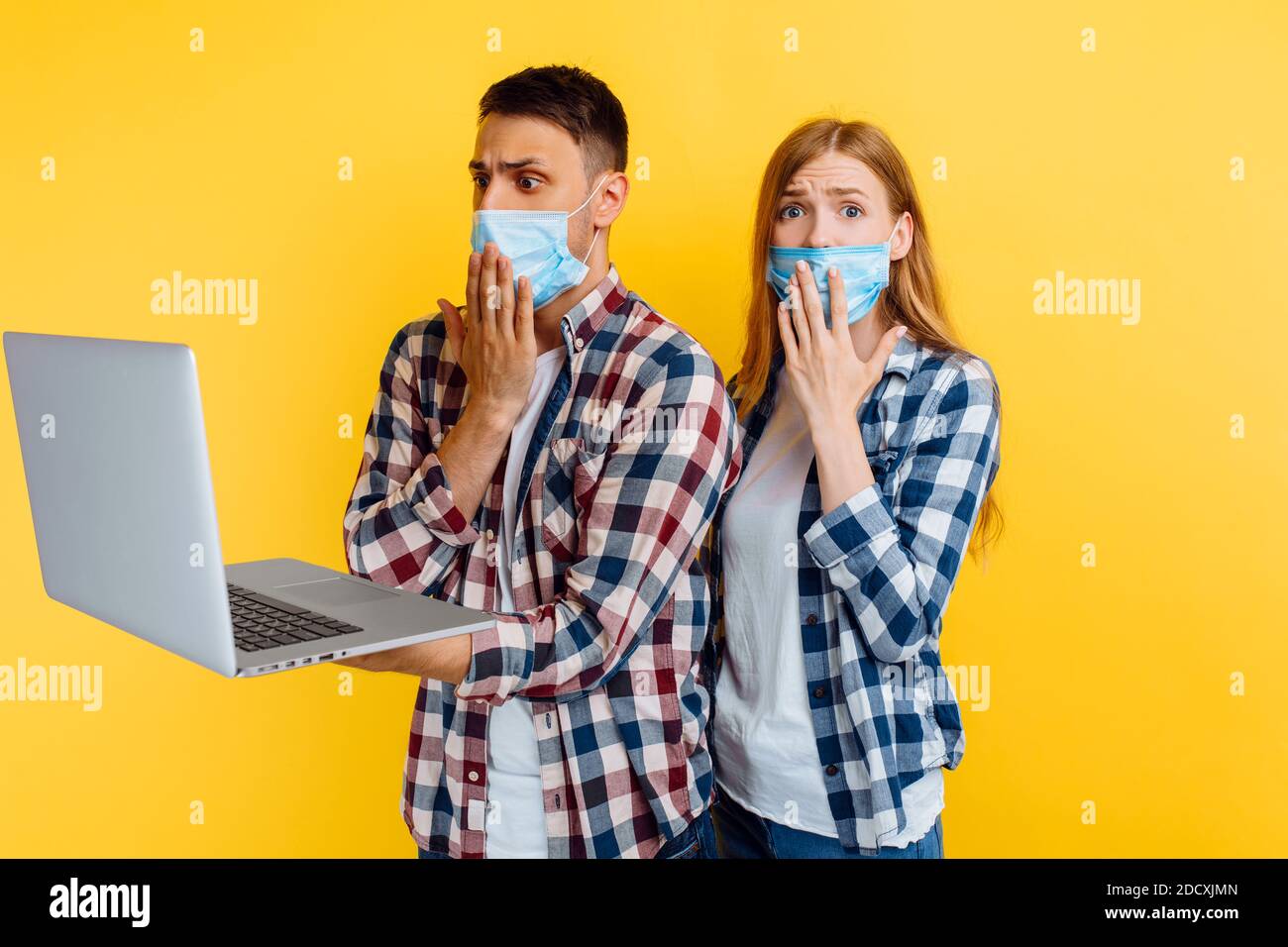 shocked man and woman in plaid shirts and medical protective masks on their faces, working on a laptop computer, looking at the laptop screen in surpr Stock Photo