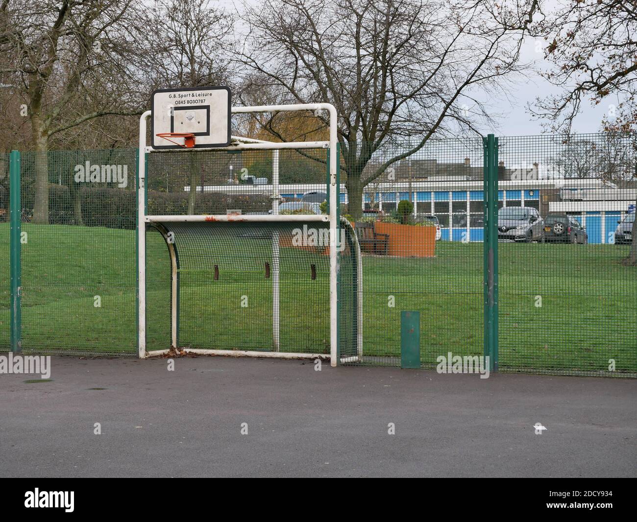 Orange basketball hoop white metal goal posts combined hard playing surface with tall green fencing surround grass behind Stock Photo