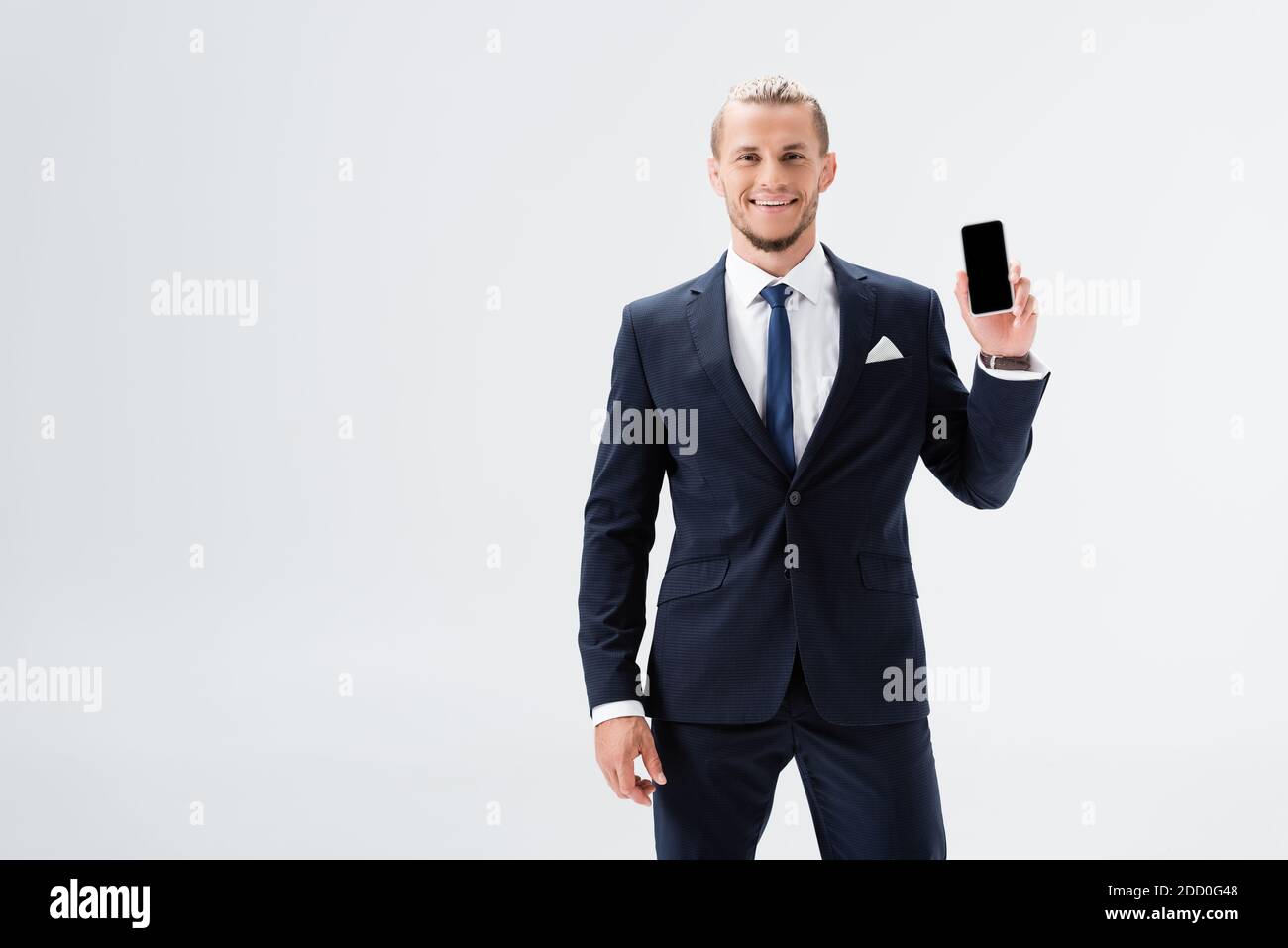 smiling young businessman in suit presenting smartphone isolated on ...