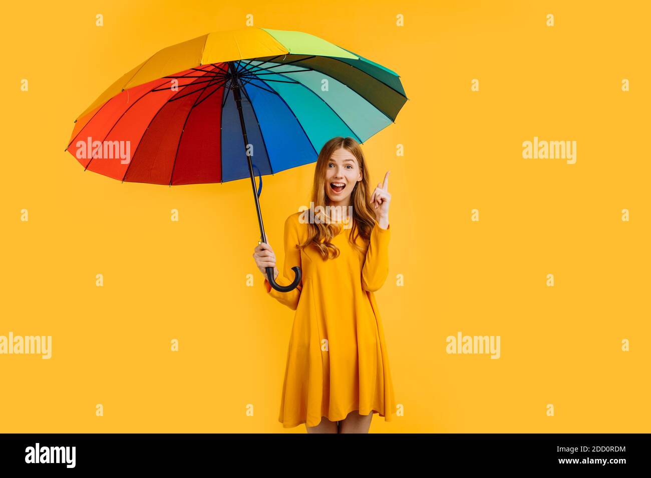 shocked girl in a yellow dress, holding a rainbow umbrella, points to an empty space for a copy of the space on a yellow background Stock Photo