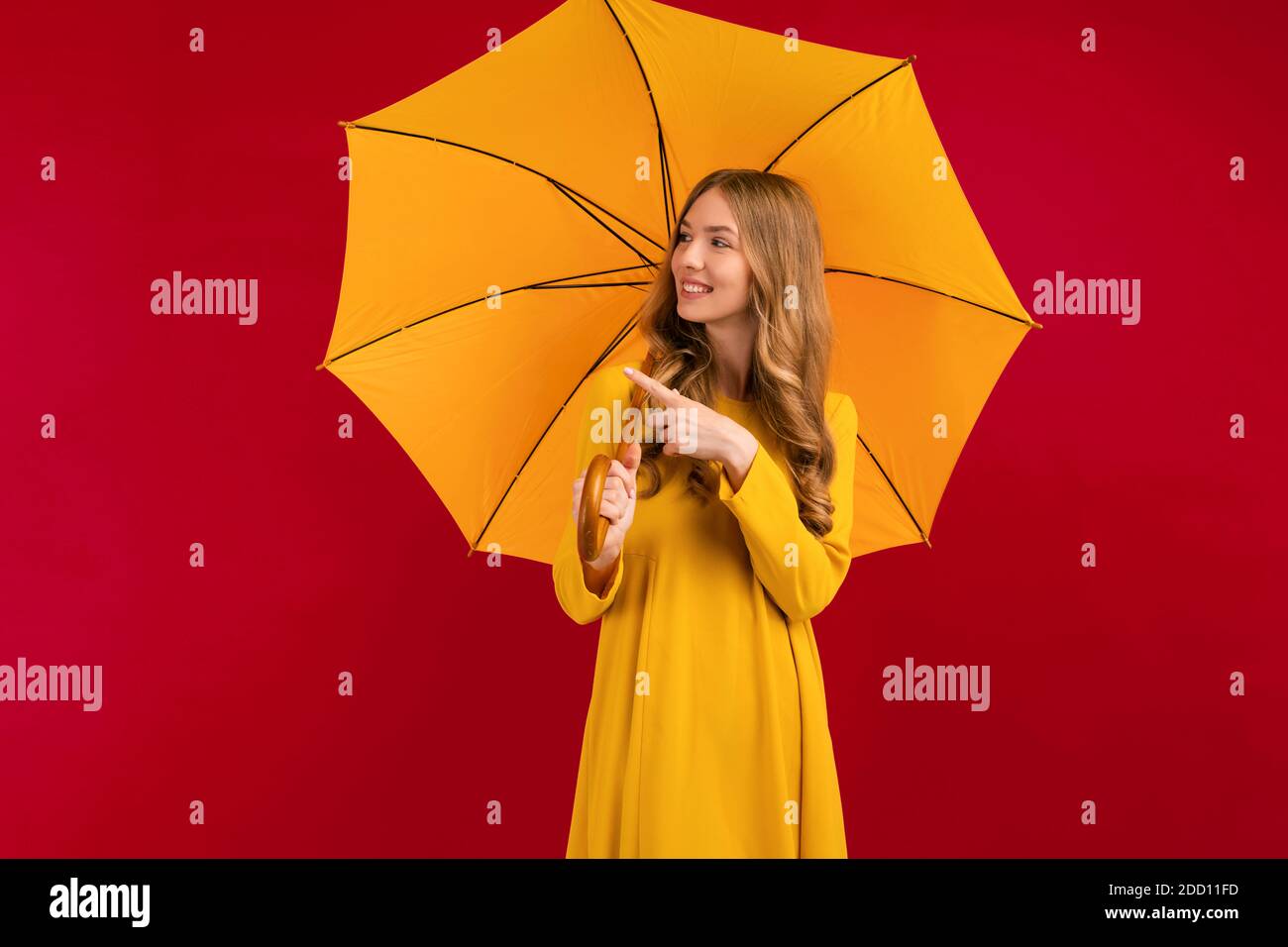 happy young woman with a yellow umbrella pointing at a copy of the space on an empty red background, autumn concept Stock Photo