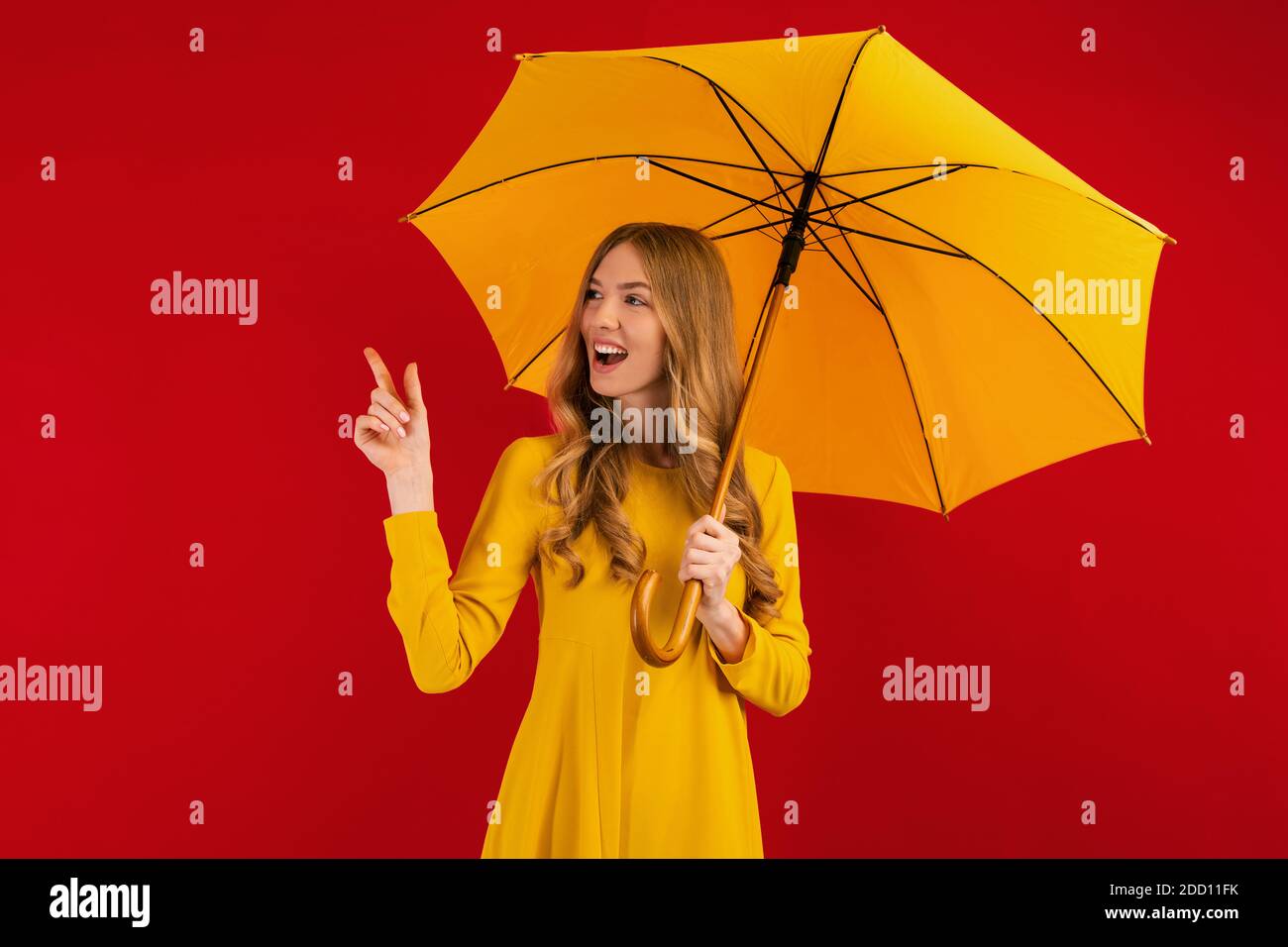 happy young woman with a yellow umbrella pointing at a copy of the space on an empty red background Stock Photo