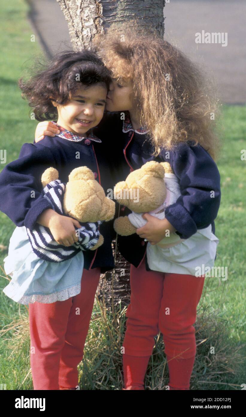 two little sisters standing under tree with teddies, big sister whispering into little one's ear Stock Photo